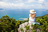 Japanese woman wearing hat sitting on rock on a cliff, ocean in the background.
