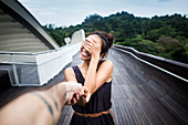 Smiling young woman standing on a bridge, covering her face, holding man's hand.