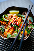 High angle close up of chopsticks on bowl with Asian food containing noodles, prawns, vegetables and chili garnish.