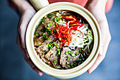 High angle close up of hands holding bowl with Asian soup containing rice vermicelli, beef and chili garnish.