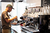 Bearded man wearing baseball cap standing at espresso machine a restaurant.