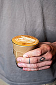 Close up of person with tattooed finger holding glass of cafe latte.