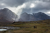 KAZAKH RIDERS BENEATH THE RAYS OF LIGHT IN CLOUDY WEATHER, REDDISH HILLS WITH SNOW-COVERED SUMMITS IN THE DISTANCE, TAVAN BOGD MASSIF, ALTAI, BAYAN-OLGII PROVINCE, MONGOLIA