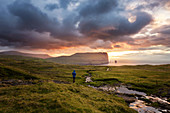 Wanderer auf einer grünen Fläche stehend betrachtet den bunten Sonnenuntergang vor den großen Meeresklippen, Eidi, Eysturoy, Färöer, Dänemark