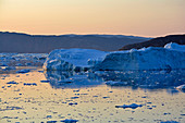 Fjord on the Eqi Glacier in West Greenland; Icebergs and ice floes in calm water; Mountains in the background; Evening sun colors sky and water surface light orange; Dark sides of the icebergs and floes glow blue