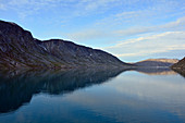 Southwestern Greenland; Entering the Ikka Fjord; steep mountain slopes frame the fjord; early morning; the sun is still behind the mountains; Rocks and debris; little vegetation; nice weather with light clouds; Mountains and clouds are reflected on the water surface