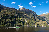 Ausflugsboot gleitet über das glatte Wasser vor Panoramalandschaft aus hohen, schroffen Bergen und Wasserfällen, Milford Sound, Fiordland National Park, Südinsel, Neuseeland