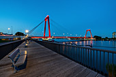 Illuminated, red Willemsbrücke during the blue hour, with plank path and seating on the river bank. Rotterdam, The Netherlands, June 2020