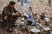 Man sitting by a camp fire in a forest, boiling kettle of water.