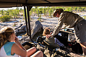 A jeep with two children and a tour guide leaning out and looking at the carcass of a dead elephant.