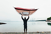 Man holding camping tent over head, standing on rocky beach,an inlet on the Alaska coastline.