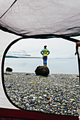 View through camping tent doorway of woman standing on beach,an inlet on the Alaska coastline.
