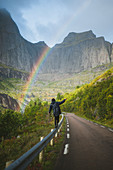 Norwegen, Lofoten, Mann balanciert auf Leitplanke mit Bergen und Regenbogen im Hintergrund