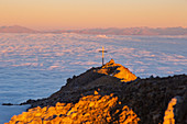 Summit cross of Latemar mount at sunrise over a cloudy carpet during summer. Schenon mount, Latemar group, Costalunga pass, Fassa valley, Pozza di Fassa, Trento district, Dolomites, Italy, Europe.