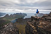HIKER  ON A PROMONTORY AT THE SUMMIT OF THE GRYTETIPPEN ABOVE THE FJORDS AND MOUNTAINS OF THE ISLAND OF SENJA, NORWAY