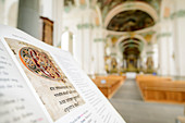 Geschmückte Seite eines mittelalterlichen Buches mit Altarraum unscharf im Hintergrund, Stiftskirche St. Gallen, St. Gallen, UNESCO Weltkulturerbe St. Gallen, Schweiz