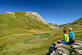Man and woman while hiking sit on rock spur and look at pastureland and mountains, at Col de Peyrelue, Pyrenees National Park, Pyrénées-Atlantiques, Pyrenees, France