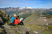 Man and woman while hiking sit at break and look at map, Großvenediger in the background, Granatspitzgruppe, Hohe Tauern, Hohe Tauern National Park, East Tyrol, Austria