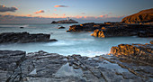 Sonnenuntergang über dem Godrevy-Leuchtturm auf der Insel Godrevy in der St Ives Bay mit Strand und Felsen im Vordergrund, Cornwall, Großbritannien