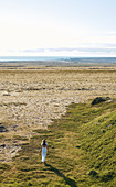 Eine Frau in einem langen weißen Kleid, die von der Kamera entlang eines Grasstreifens in Richtung Sandstrand und Meer weggeht.