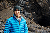 Ein Mann, der einen blauen gepolsterten Mantel trägt, der die Kamera mit einem grauen steinigen Hügel im Hintergrund betrachtet.
