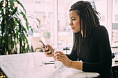 Young woman with dreadlocks, wearing black T-Shirt sitting at table in a bar, using mobile phone.
