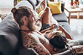 Bearded tattooed man with long brunette hair and woman with long brown hair cuddling on a sofa.
