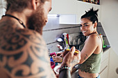 Bearded tattooed man with long brunette hair and woman with long brown hair standing in a kitchen, smiling at each other.