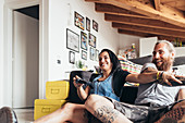 Bearded tattooed man with long brunette hair and woman with long brown hair sitting on a sofa, smiling while playing console game.