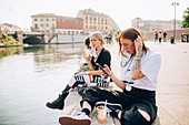 Three young women sitting on a riverbank, drinking and using mobile phone.