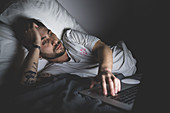 Bearded young man lying in bed at night, looking at laptop.