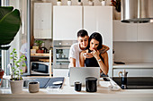 Young lesbian couple standing in kitchen, using laptop.
