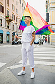 Young lesbian woman standing on a street, waving rainbow flag.