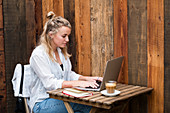 Junge blonde Frau sitzt allein an einem Cafétisch mit einem Laptop, arbeitet aus der Ferne
