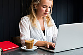Junge blone Frau mit Gesichtsmaske sitzt allein an einem Cafétisch mit einem Laptop, arbeitet aus der Ferne