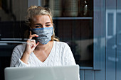 Young blond woman wearing face mask sitting alone at a cafe table with a laptop, using mobile phone, working remotely.