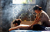 Ein balinesischer Therapeut, der eine Massage an einer asiatischen Frau in einer rauchigen, romantischen Umgebung durchführt. Bali, Indonesien.