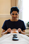 Eine Spa-Therapeutin führt eine Kopfmassage durch, während der Gast heiße Steine auf ihrem Körper genießt. Bath, Vereinigtes Königreich