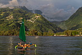 Menschen genießen Törn auf einem Auslegerkanu mit Segel vor üppiger Bergkulisse, nahe Papeete, Tahiti, Windward Islands, Französisch-Polynesien, Südpazifik