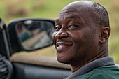 Porträt eines lächelnden ruandischen Mannes in einem Safari Fahrzeug betrieben vom Luxusresort Zeltcamp Magashi Camp (Wilderness Safaris), Akagera National Park, Eastern Province, Ruanda, Afrika