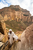 Ethiopian priest leaning on steep rocks leading to Abuna Yemata Guh church, Gheralta Mountains, Tigray Region, Ethiopia, Africa
