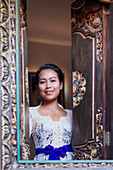 A Balinese woman in traditional dress looking out from a carved window in a local village, Bali, Indonesia, Southeast Asia, Asia