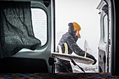 Woman with a black jacket and woolly hat putting a surfboard into the back of a campervan. Winter Surfing