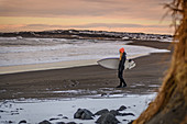 A woman standing and holding a surfboard on a beach looking out to sea.