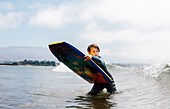 Portrait of young boy wearing wet suit in ocean, holding bodyboard, waiting for wave, Santa Barbara, California, USA.