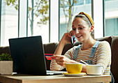 Woman seated in a cafe using a laptop and speaking on a smart phone, holding a credit card