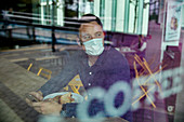 Man in a face mask seated at a cafe table, view through a window