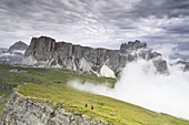Hikers descend from Mount Mondeval while fast and dense clouds rise from the valley, in the center the Lastoi Di Formin, in the background on the left the Tofana di Rozes
