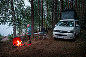 Frau in Outdoorkleidung bei Vanlife Camping mit VW Bus im Wald in Schweden, am See mit Lagerfeuer, Siljansee
