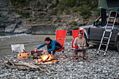 Albania, Southern Europe, young couple sitting in front of off-road vehicle with roof tent, campfire, river, Vjosa, Permet